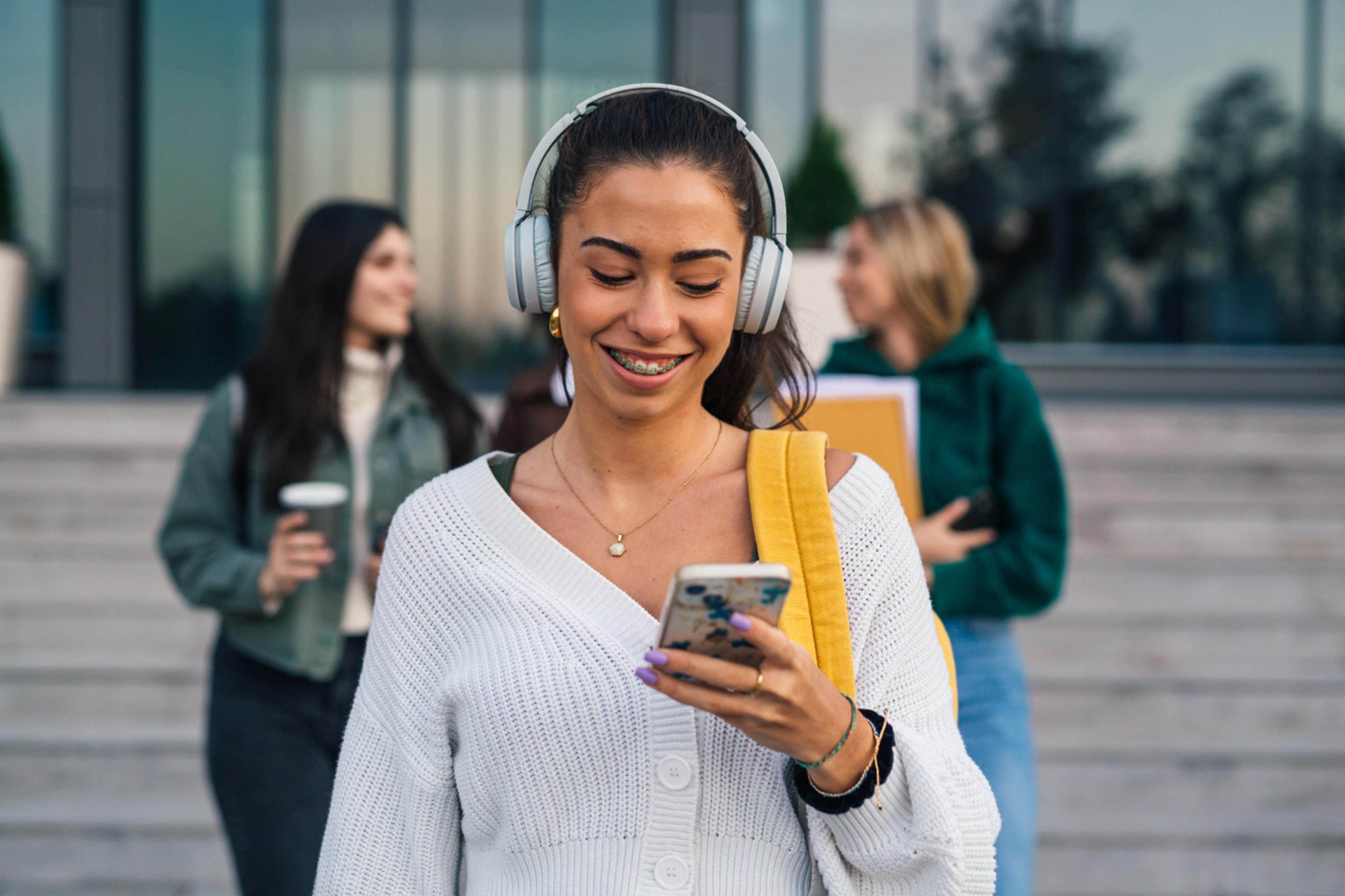 smiling college student wearing headphones while looking at cell phone