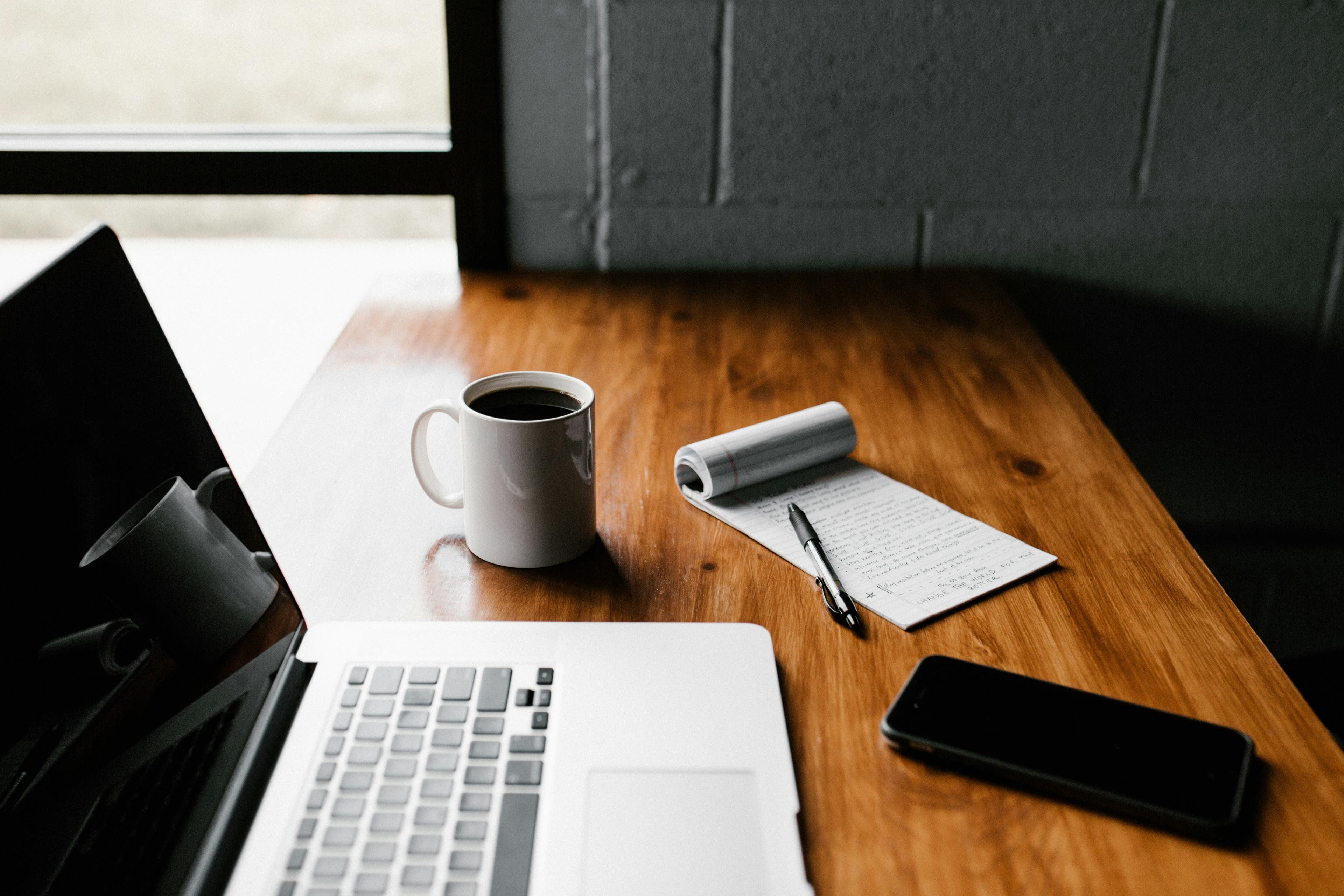 A computer, coffee cup, and notebook sit on a desk in a well-lit workspace