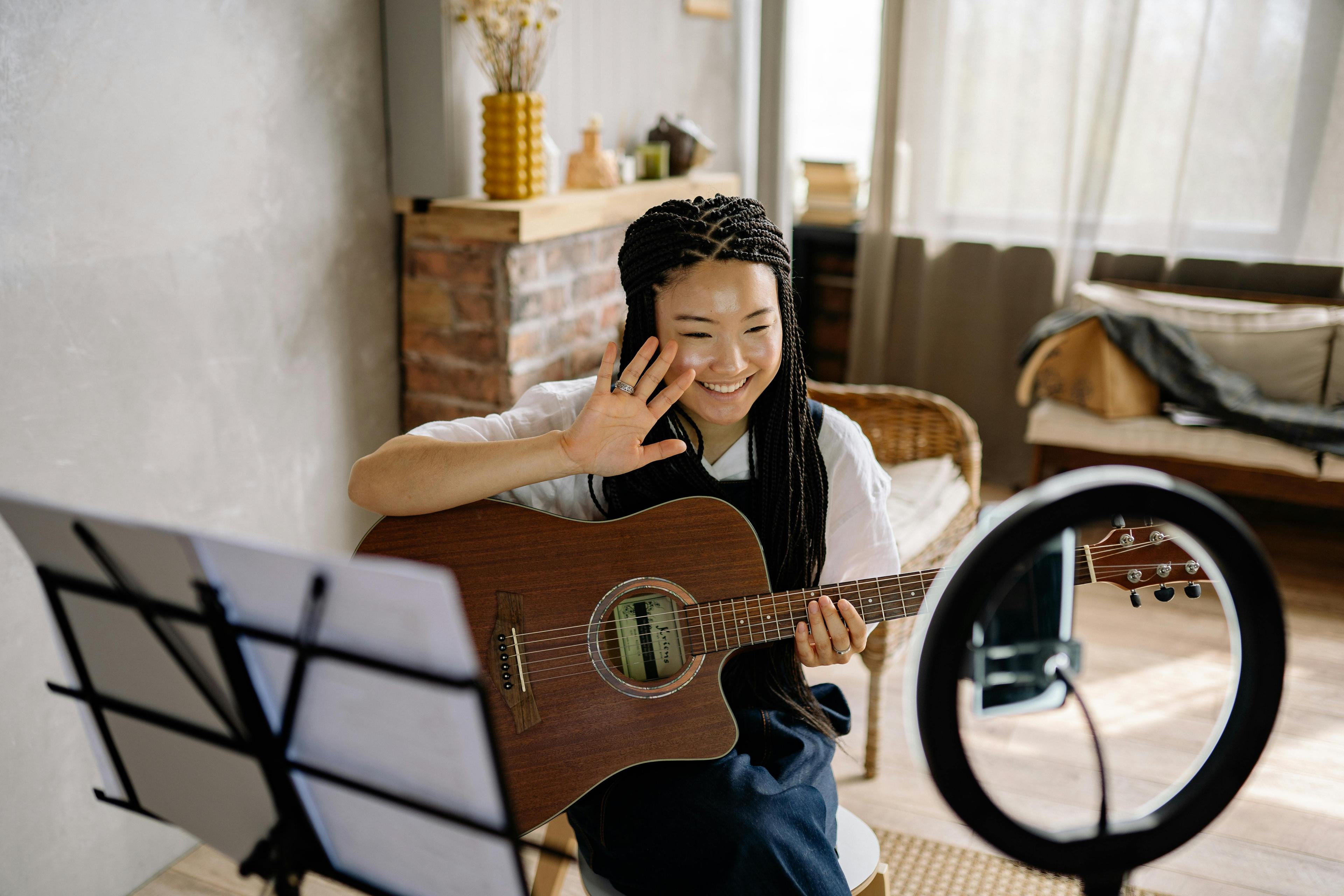 Young content creator holds a guitar and waves to a mobile phone set up on a ring light tripod