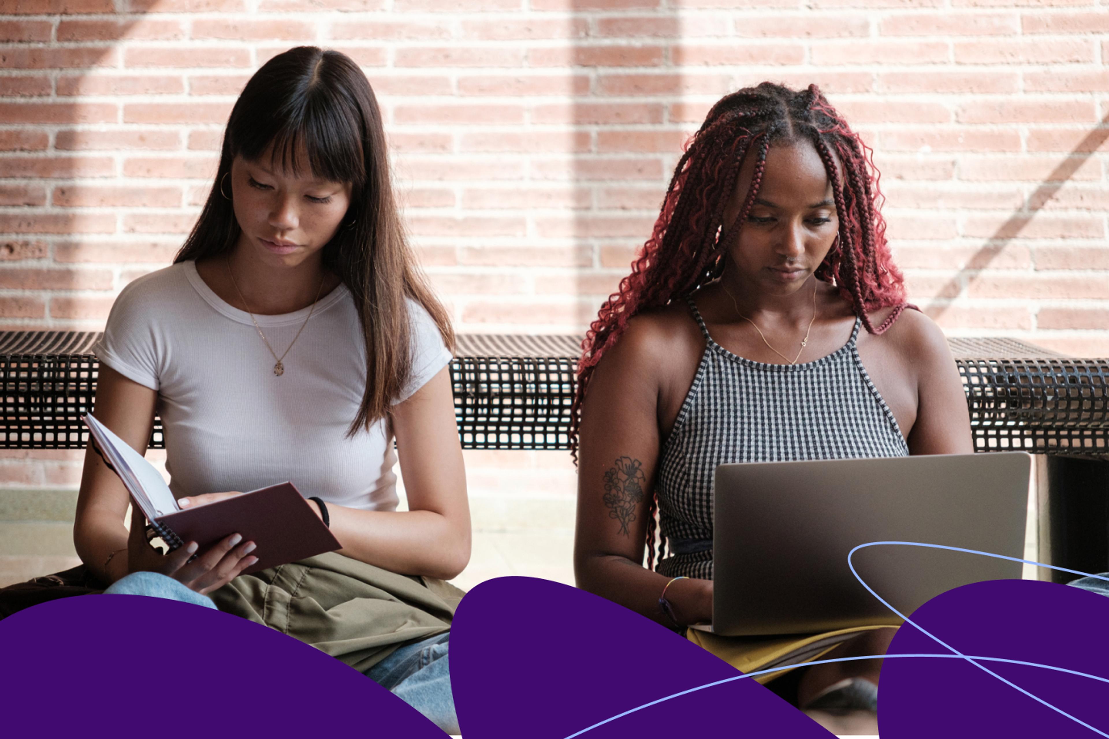 Two women on a bench, one looking at a book, one looking at a laptop