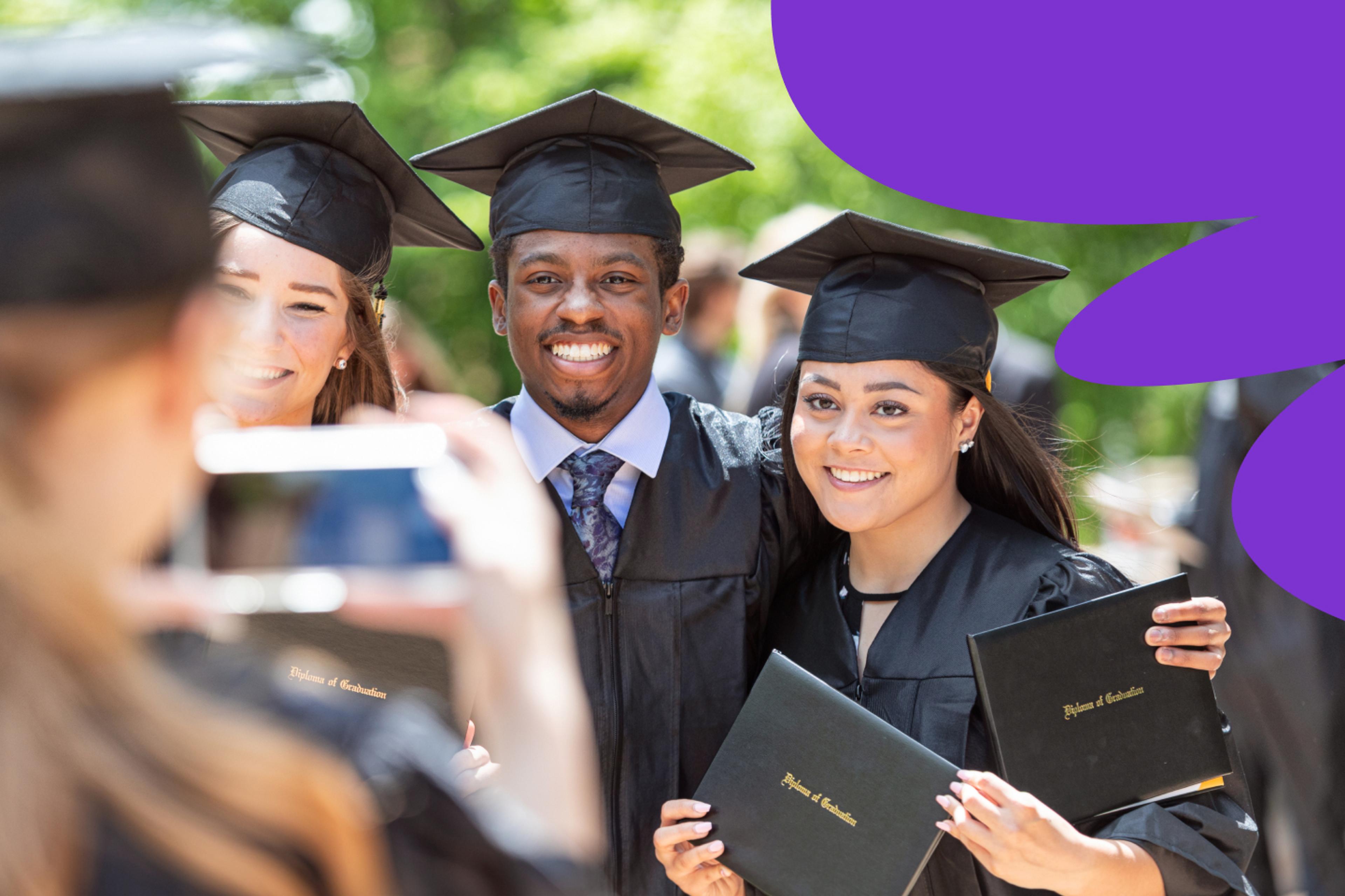 Three graduates in cap and gown smiling for a photo holding up their diplomas